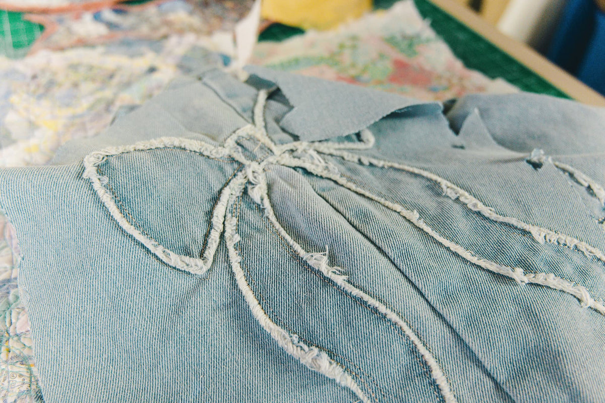 A close-up of a light blue denim garment featuring a large bow design outlined with distressed white embroidery, resting on a patterned surface.