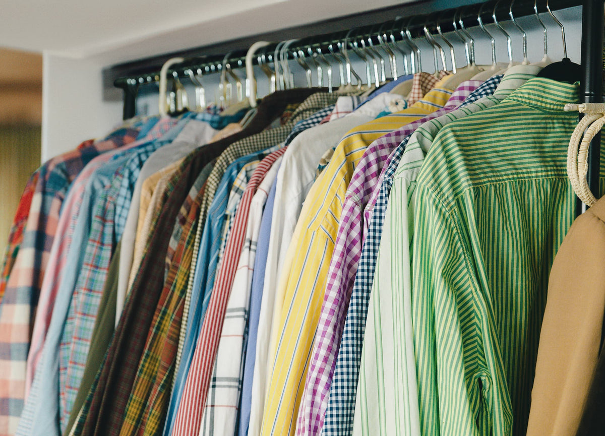 A row of colorful long-sleeve button-up shirts hanging on white hangers from a black rail, with a green striped shirt in the foreground.