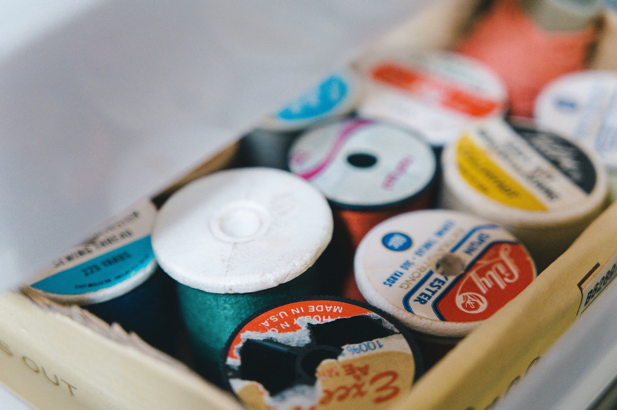 A close-up, slightly out-of-focus view of multiple vintage spools of thread stored inside a cardboard box. The spools feature various colors and labels, including a prominent teal green spool in the foreground and a red and white 'Fly' label.