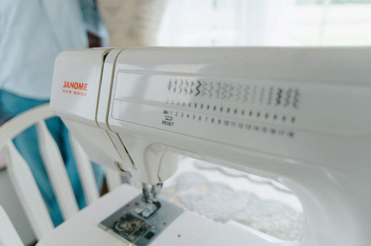A close-up of a white Janome sewing machine on a white table, with a person in a light blue shirt and jeans blurred in the background near a window.
