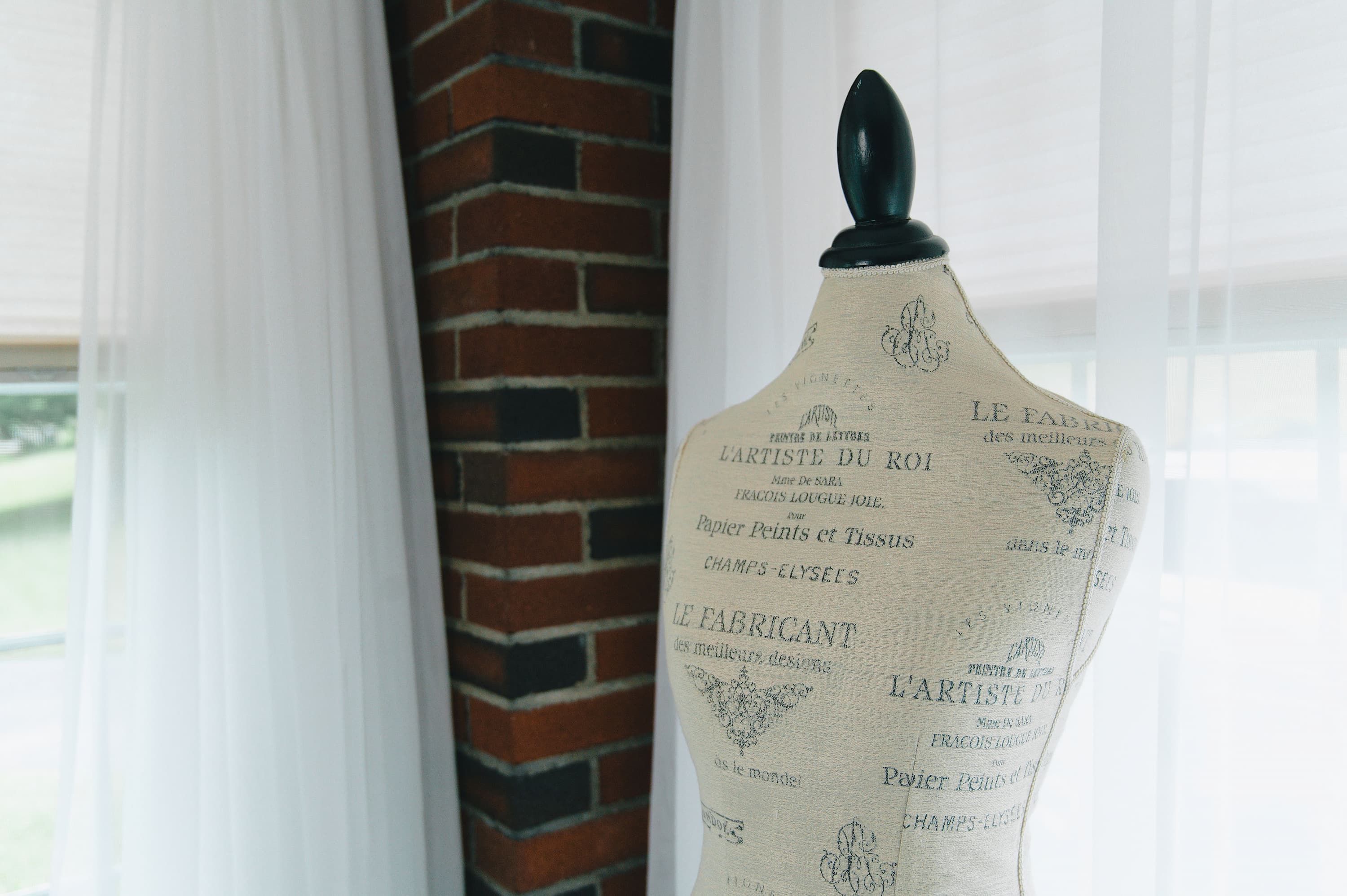 A vintage-style fabric dress form mannequin with a black neck cap, covered in beige French text, set against a background of white curtains and a red brick column.