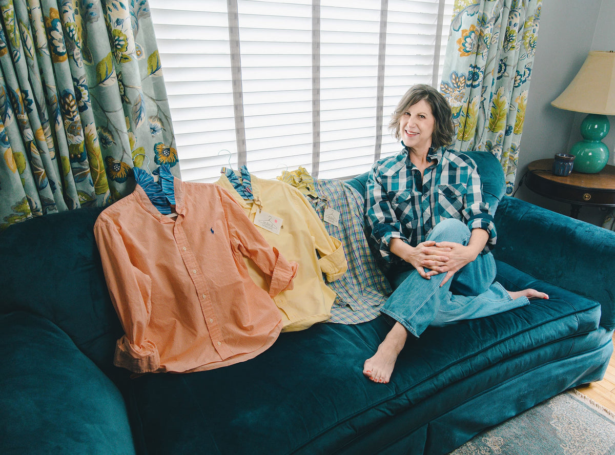 Elizabeth Gasper sits barefoot on a teal sofa in a living room, wearing a plaid shirt while several other shirts are draped over the couch. The room has floral curtains, window blinds, and a side table with a lamp.