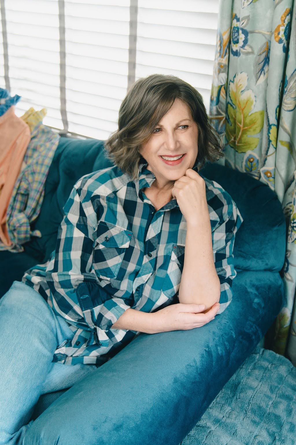 A woman with short gray-brown hair sits smiling on a teal velvet armchair in a home setting, wearing a blue and white plaid button-down shirt and light blue jeans.