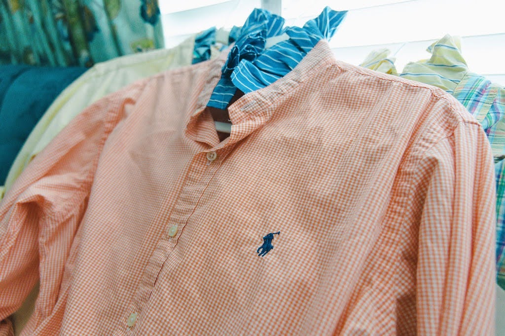 A peach and white gingham long-sleeve button-down shirt with a small navy blue embroidered polo player logo on the chest, hanging on a rack next to other shirts.