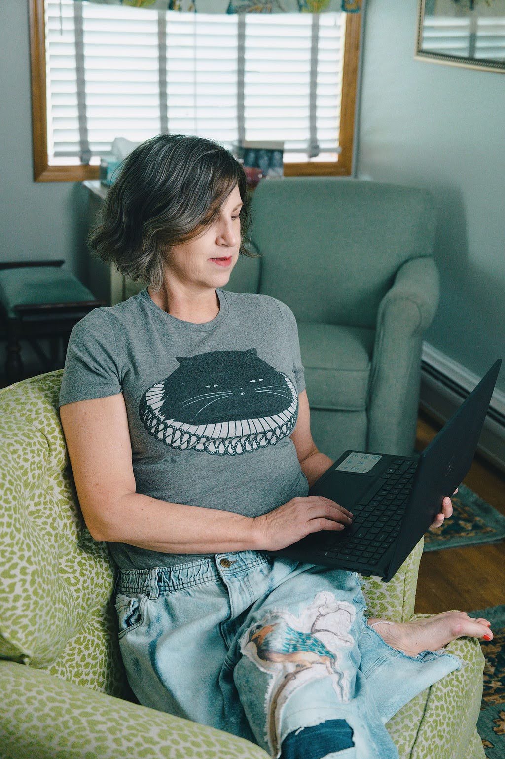 A woman sits in a patterned green armchair, looking at a black laptop she is holding. She is wearing a gray t-shirt with a black cat graphic and light blue ripped jeans in a living room setting.