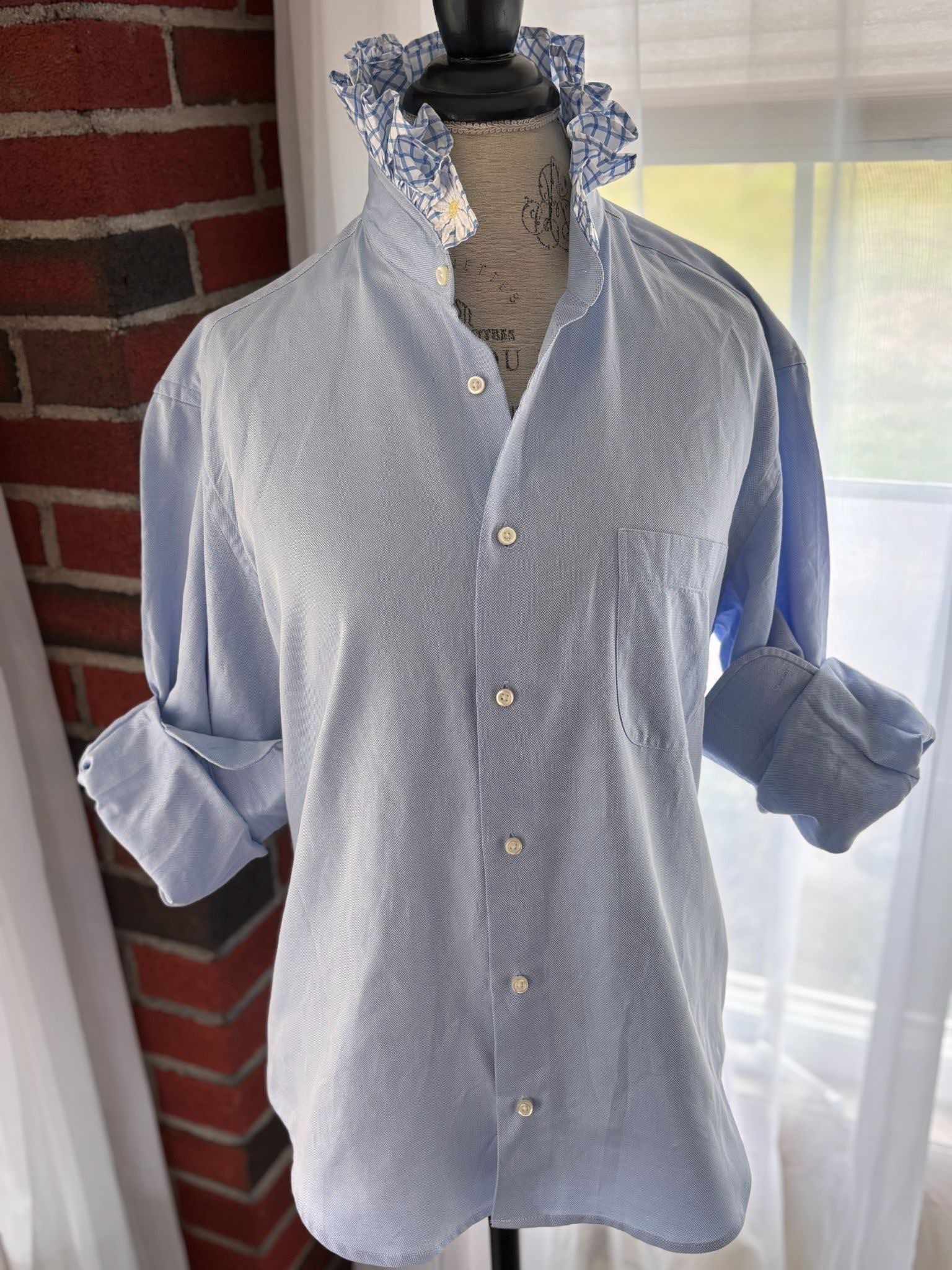 A light blue long-sleeve button-up shirt with a chest pocket, displayed on a dress form mannequin. The shirt features a pointed collar with a blue and white floral patterned lining, set against a background of a red brick wall and a window with sheer curtains.
