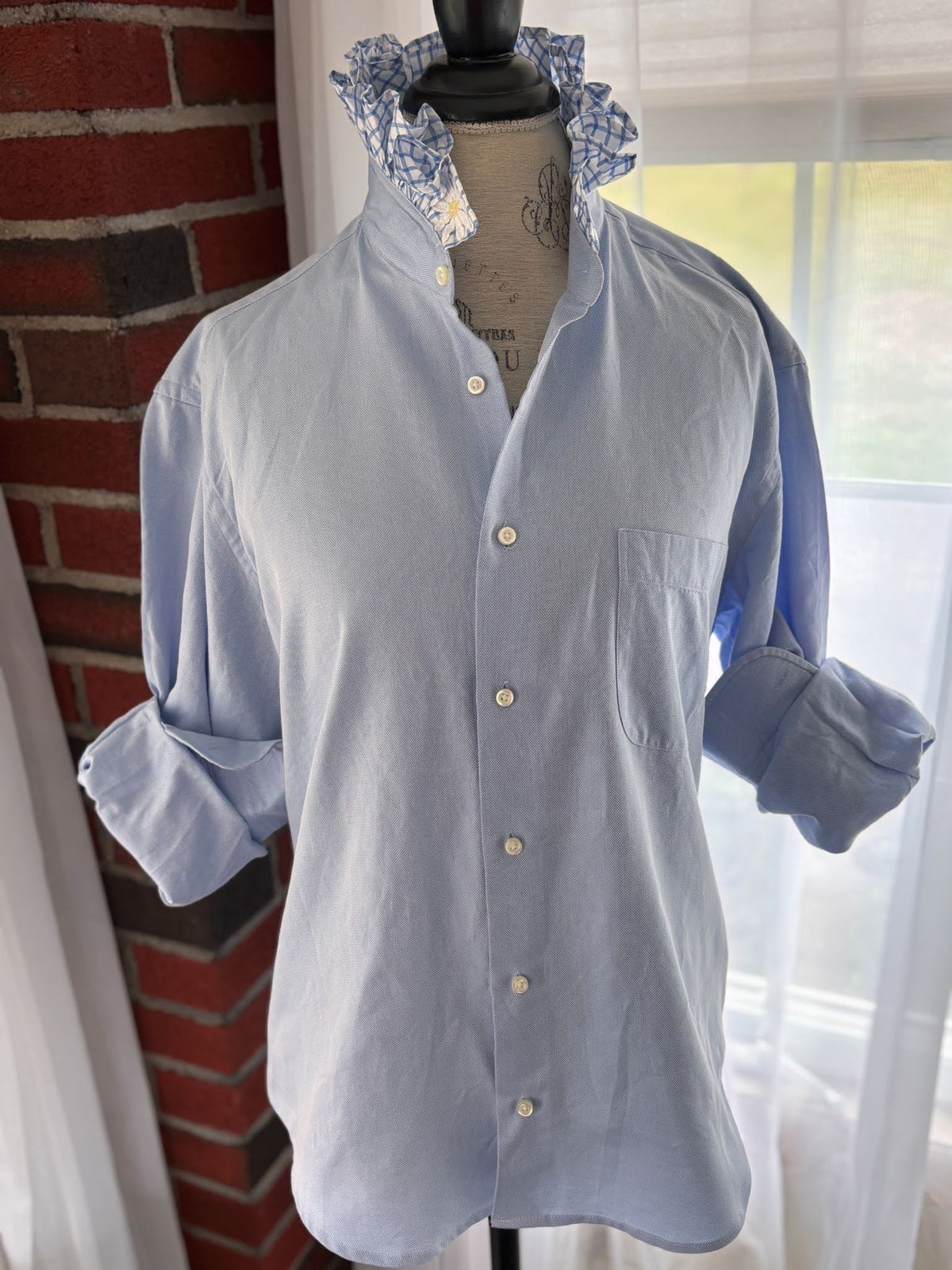 A light blue long-sleeve button-up shirt with a chest pocket, displayed on a dress form mannequin. The shirt features a pointed collar with a blue and white floral patterned lining, set against a background of a red brick wall and a window with sheer curtains.