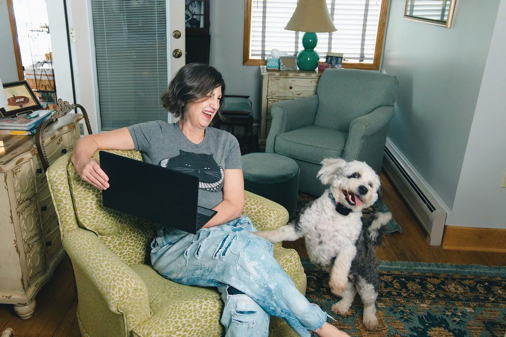 A woman sitting in a patterned green armchair with a black laptop on her lap, smiling next to a small white and gray dog in a cozy living room setting.