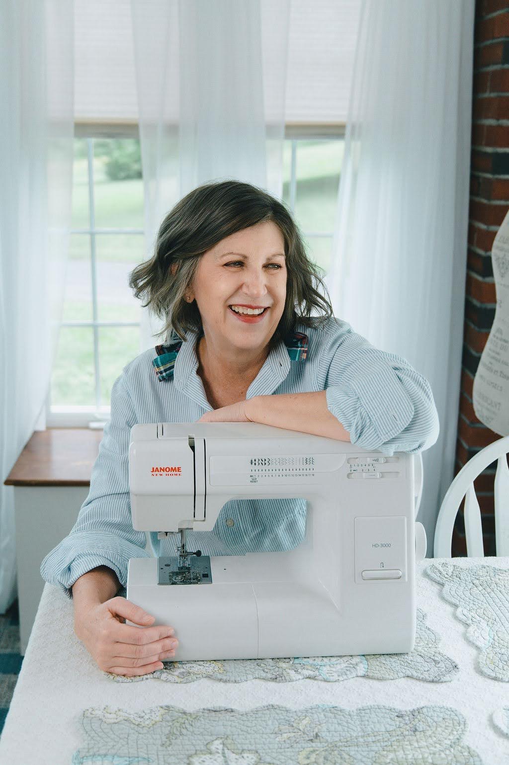 A smiling woman with short gray hair sits behind a white sewing machine on a table, with sheer curtains and a window in the background. The sewing machine has a visible brand logo.