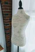A vintage-style fabric-covered dress form mannequin with a black neck cap, featuring a beige body printed with elegant French text and illustrations, set against a background of a brick column and a white curtain.