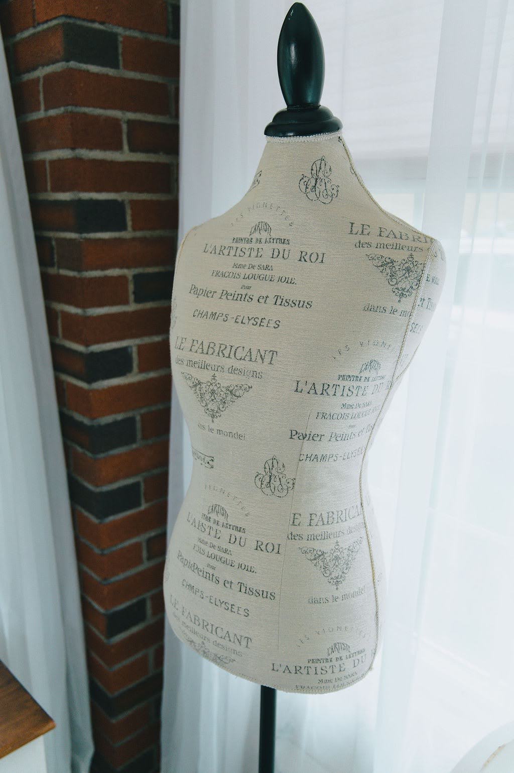 A vintage-style fabric-covered dress form mannequin with a black neck cap, featuring a beige body printed with elegant French text and illustrations, set against a background of a brick column and a white curtain.