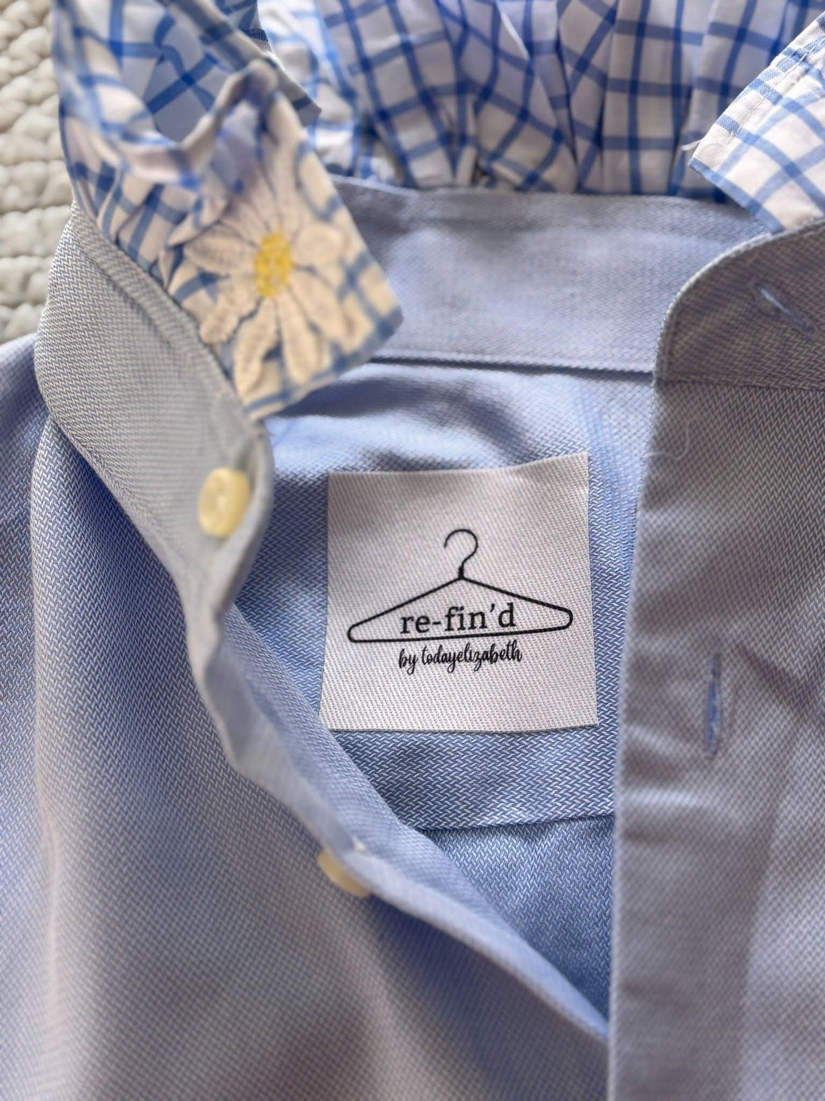 Close-up of a light blue button-down shirt with a white neck label reading 're-fin'd by todaysgents' and a blue-and-white gingham collar lining with a white daisy print.
