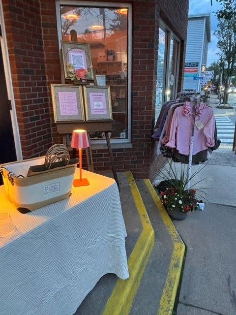 A storefront display outside a brick building featuring a white table covered with a textured white cloth, a small orange lamp, framed signs, and a clothing rack holding pink and purple tops. A sidewalk, crosswalk, and street are visible in the background.